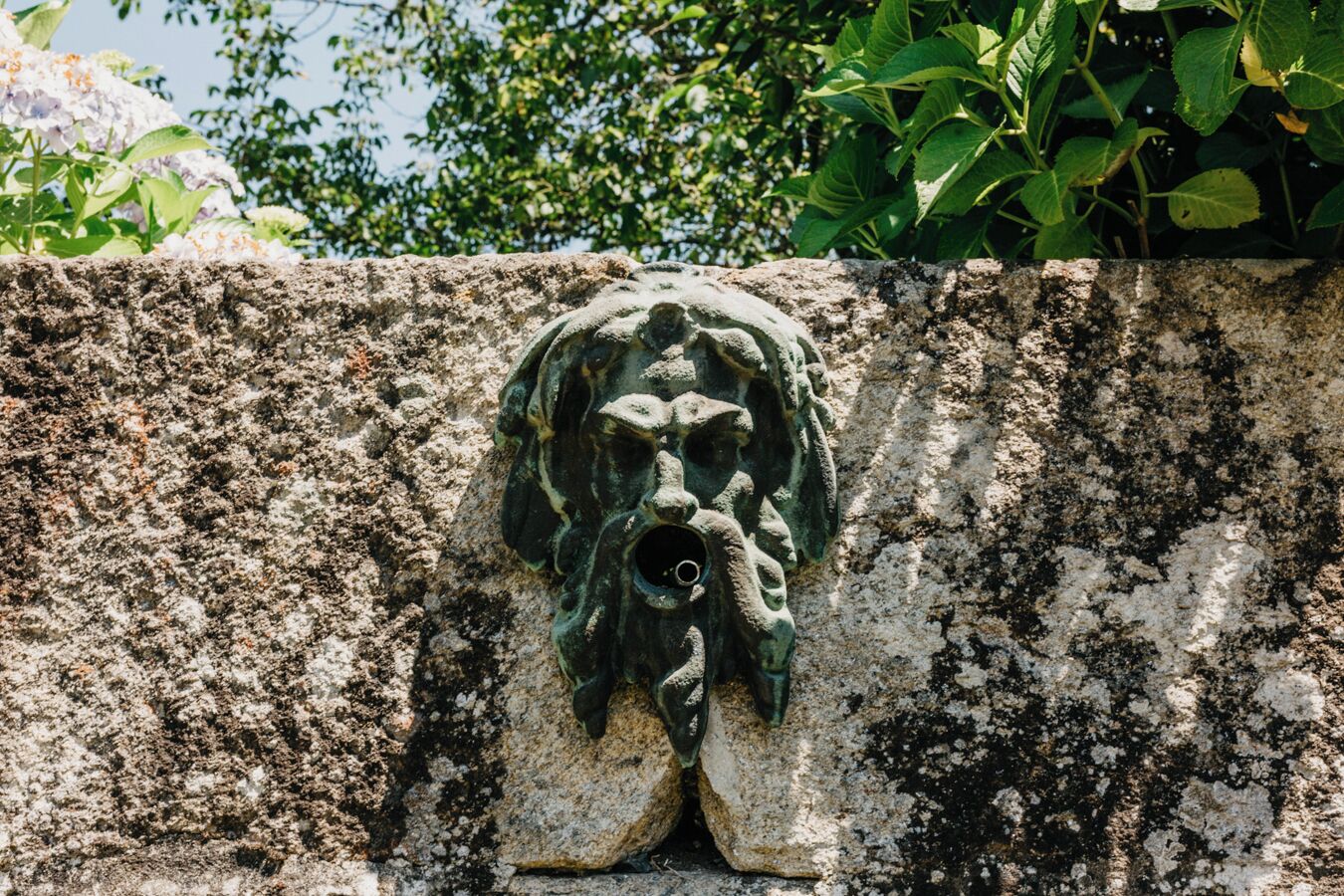 Eine Steinsäule mit einer verwitterten, grünlichen Metallskulptur eines Gesichts mit Pfeife.