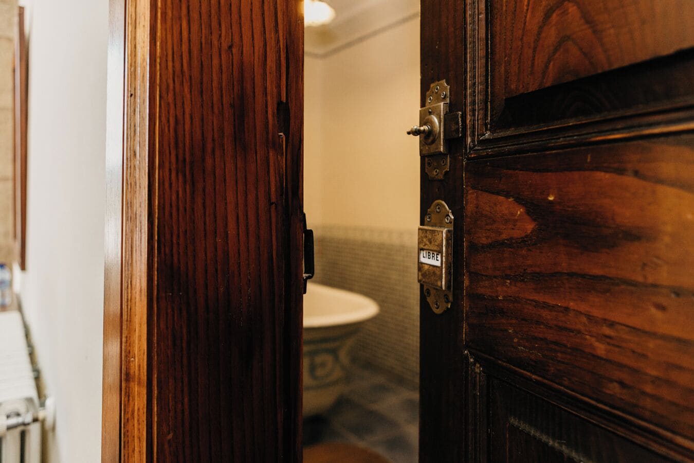 A wooden door with a brass handle and a 'LIBRE' sign, leading to a bathroom with a white toilet and a patterned floor.