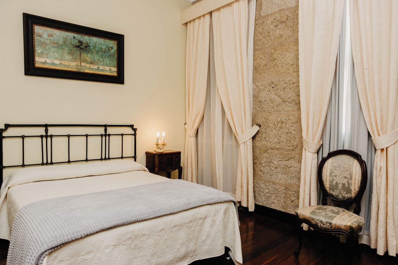 A bedroom with a bed, a nightstand, a chair, a painting, and curtains.