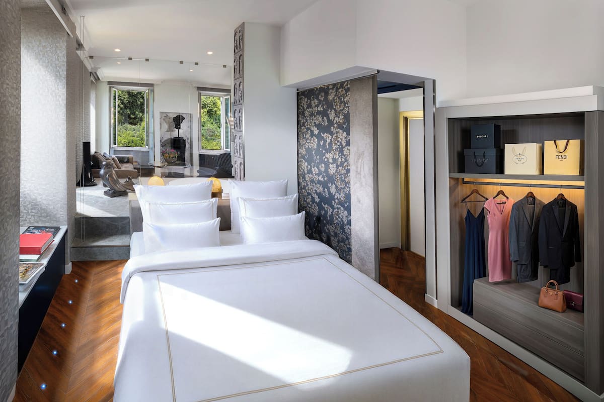 A modern hotel room with a large bed, a seating area with a sofa and chairs, a small table, and a wardrobe with clothing and bags.