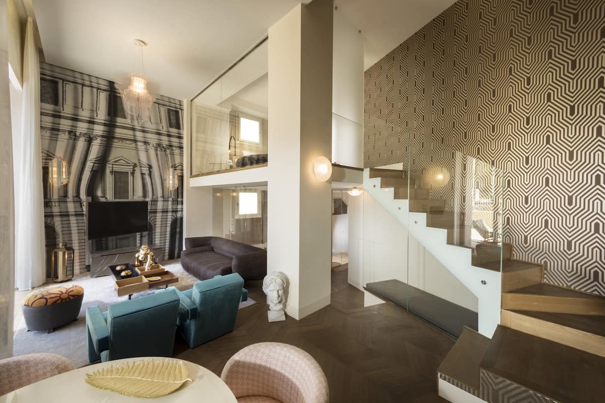 A modern living room with a sofa, a coffee table, and a TV. There is a staircase with a patterned wall and a bench.
