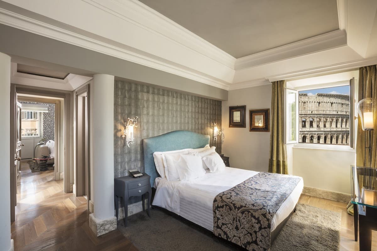 A hotel room with a large bed, a nightstand, a window with a view of the Colosseum, and a door leading to a hallway.