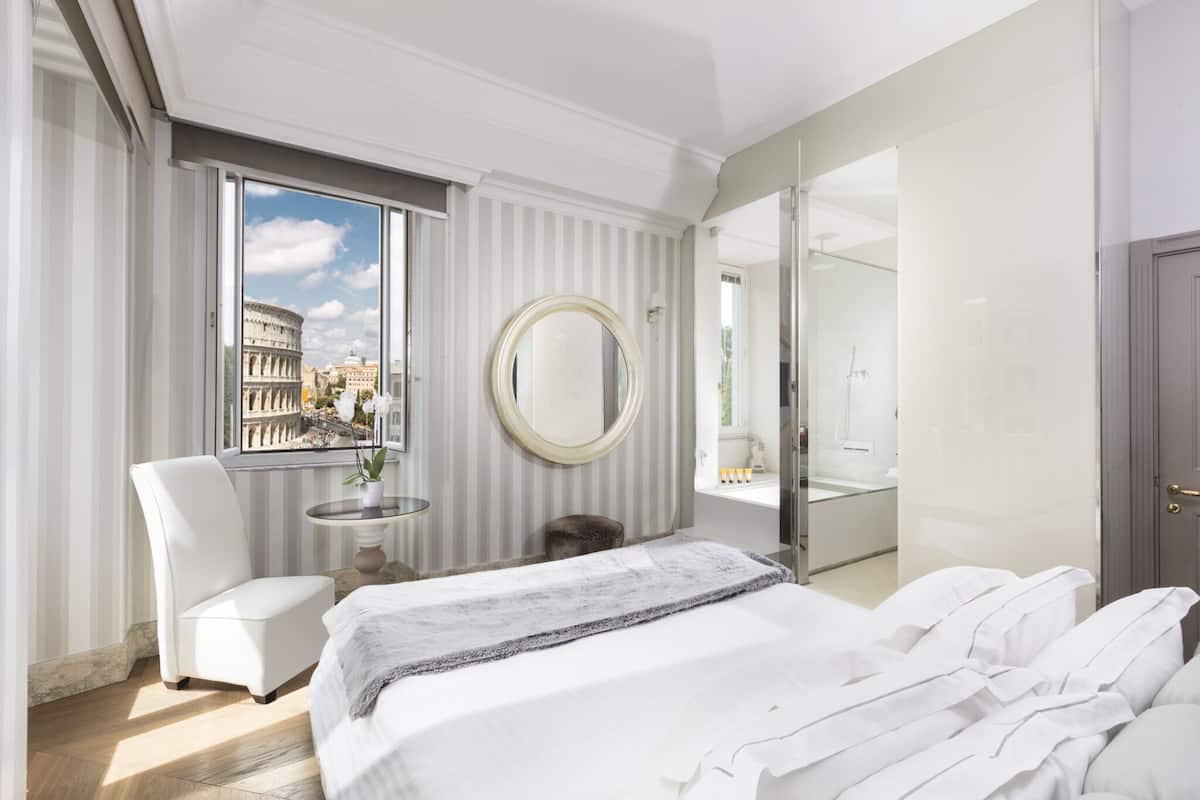 A modern hotel room with a large bed, a round mirror, a seating area with a chair, a small table with a vase, and a view of the Colosseum through a large window.