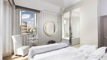 Habitación de hotel moderna con una cama grande, un espejo redondo, una zona de estar con una silla, una mesita con un jarrón y vistas al Coliseo a través de un amplio ventanal.