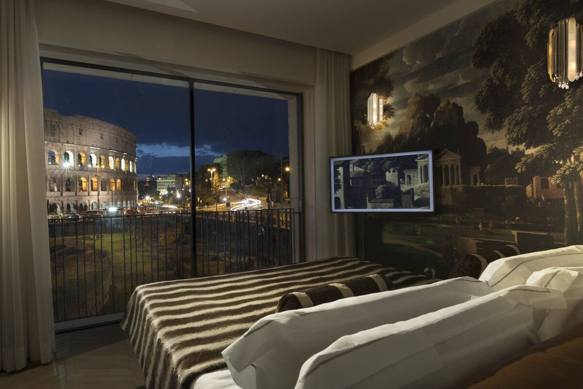 A hotel room with a large window overlooking the Colosseum at night, a bed with striped bedding, and a mural of a classical scene.