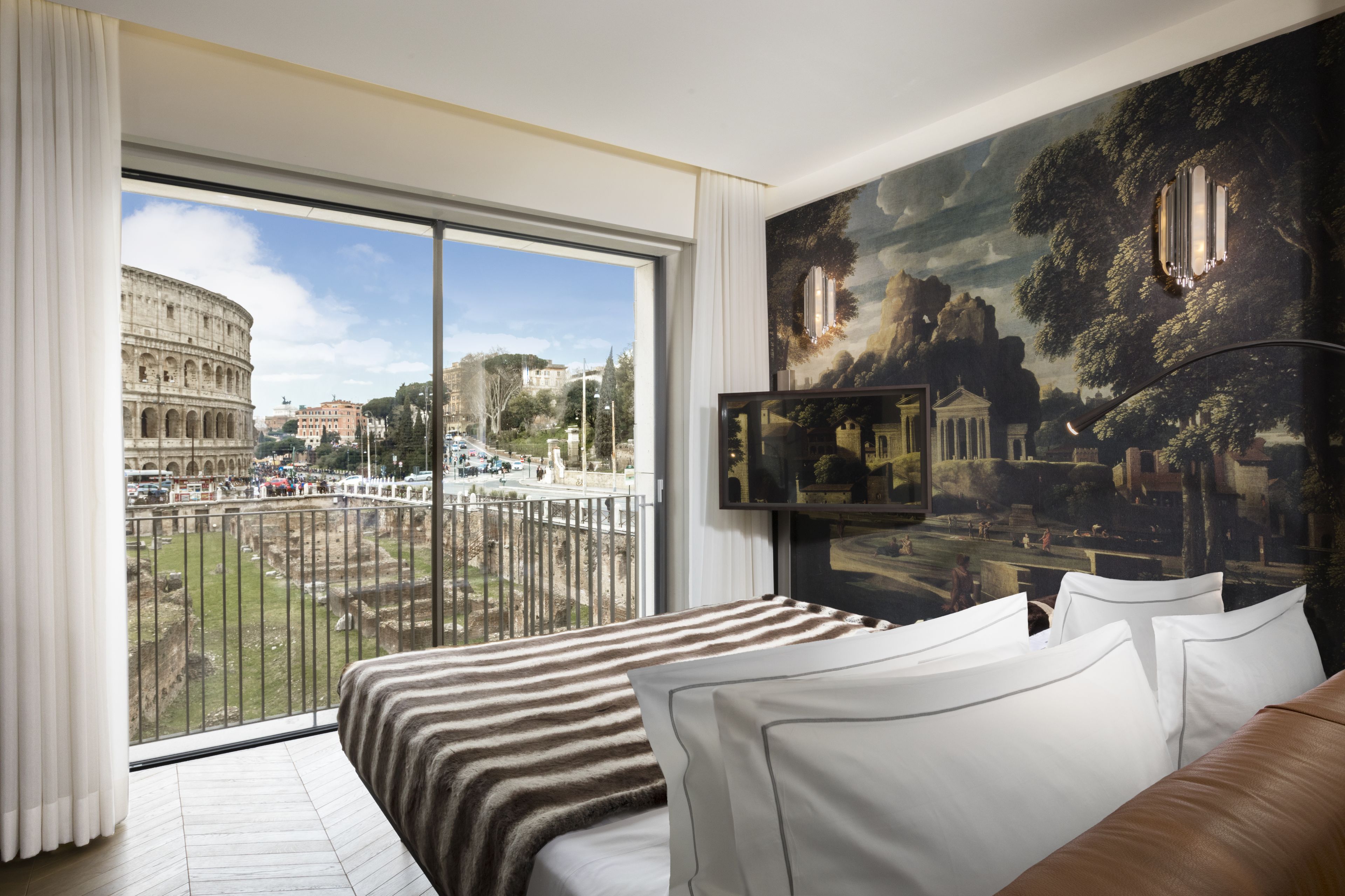 A hotel room with a large window offering a view of the Colosseum and Roman architecture. The room features a bed with striped bedding and white pillows.
