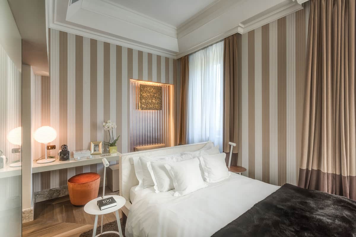 A hotel room with a bed, bedside table, lamp, and a view of the city.