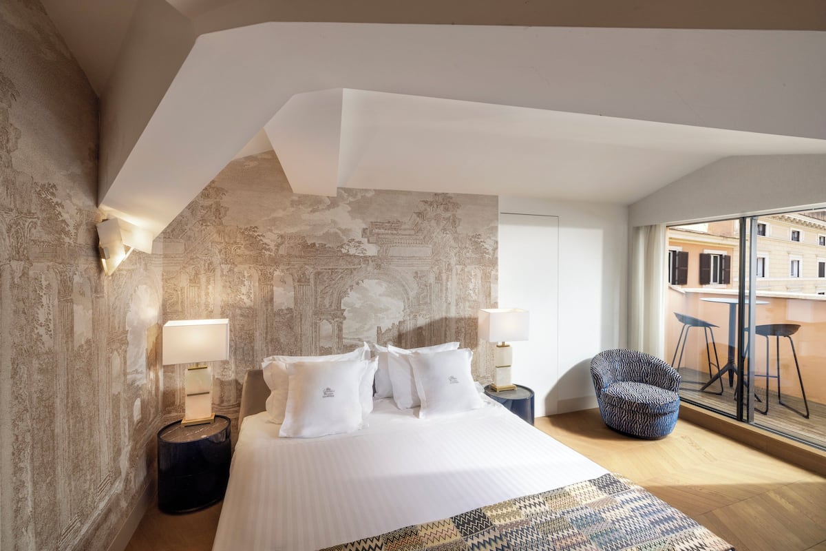 A modern hotel room with a large bed, two bedside lamps, a patterned rug, and a view of the city through large windows.
