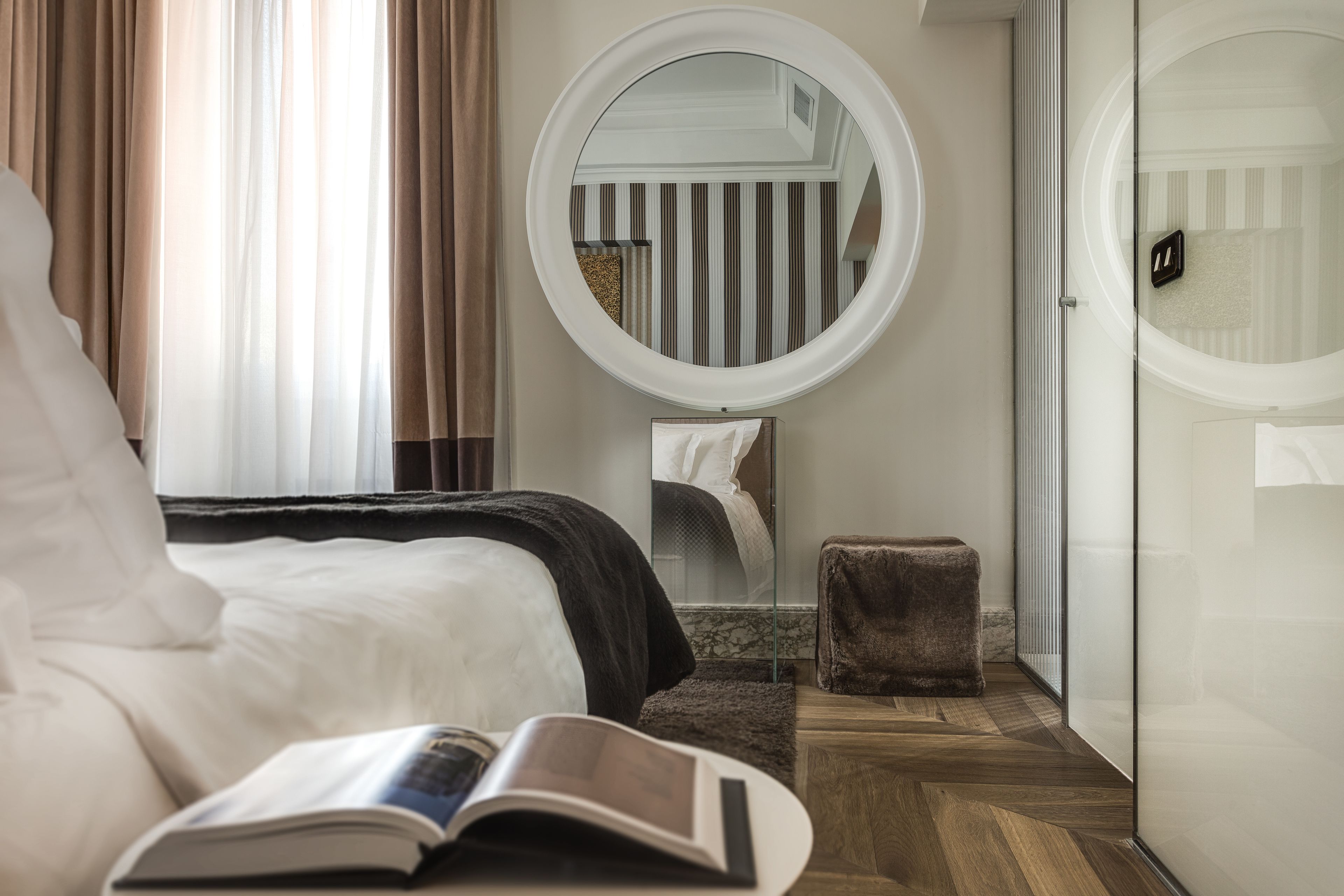 A hotel room with a bed, a round mirror, a small stool, and a book on the bed.