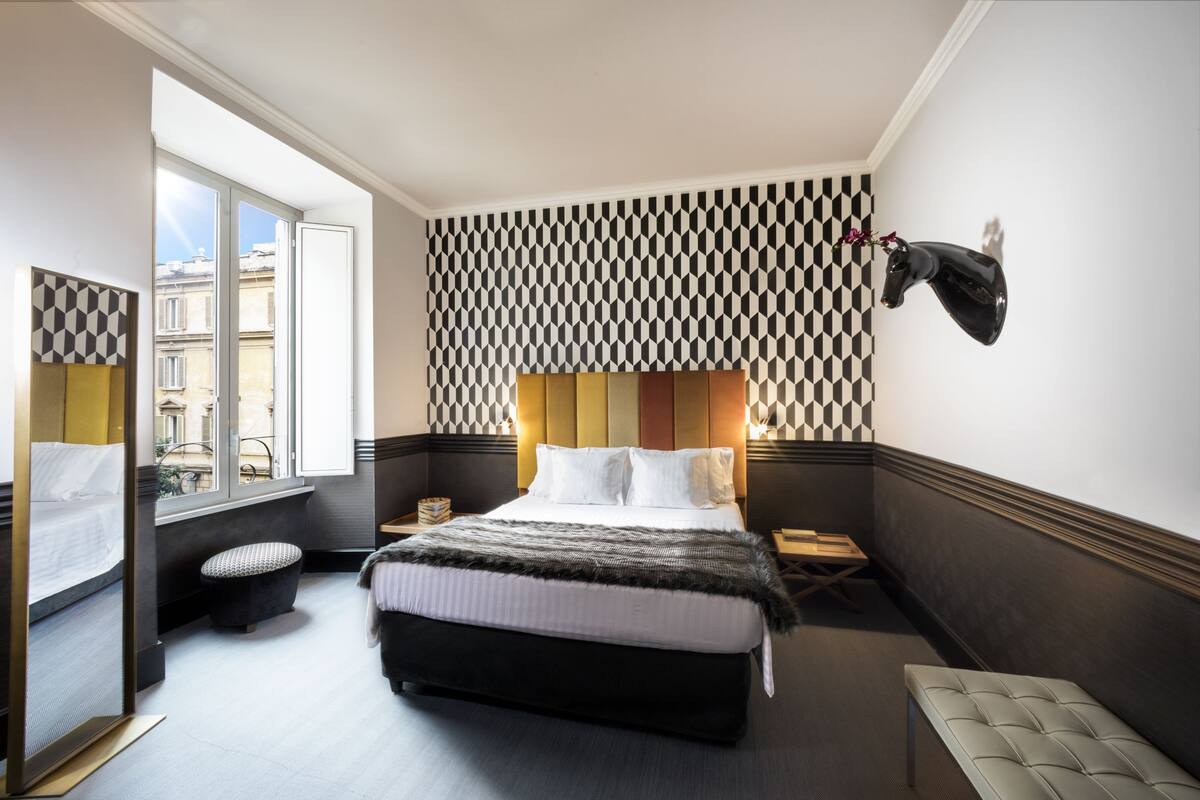 A hotel room with a large bed, a bench, a small table, a mirror, and a window with a view of buildings.