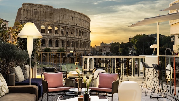 Una terraza con mobiliario al aire libre, vistas del Coliseo y un edificio moderno al fondo.
