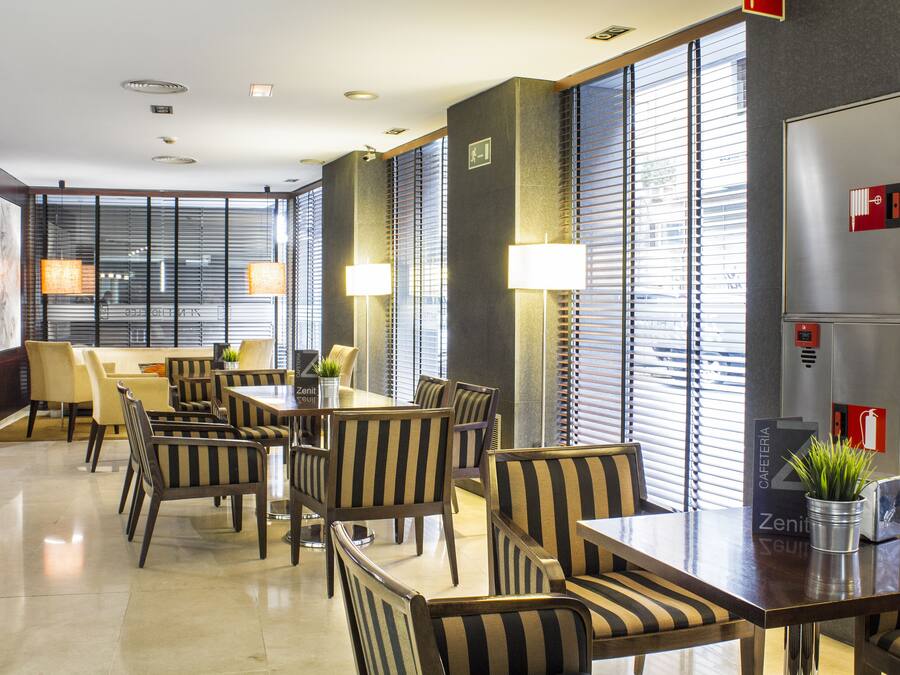 Un moderno vestíbulo de hotel con zona de comedor, sillas a rayas y grandes ventanales con persianas.