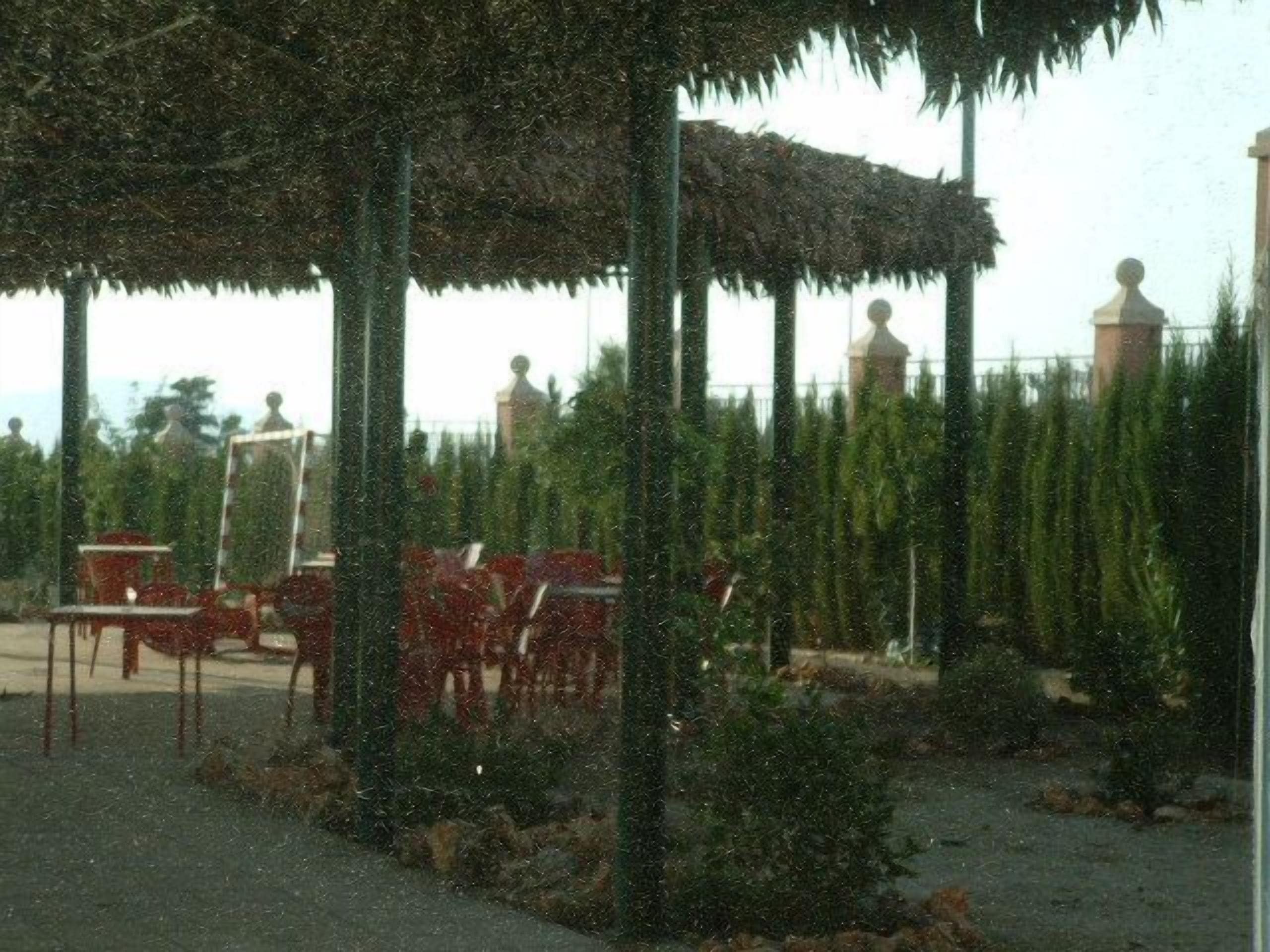 A covered outdoor seating area with tables and chairs, surrounded by greenery.