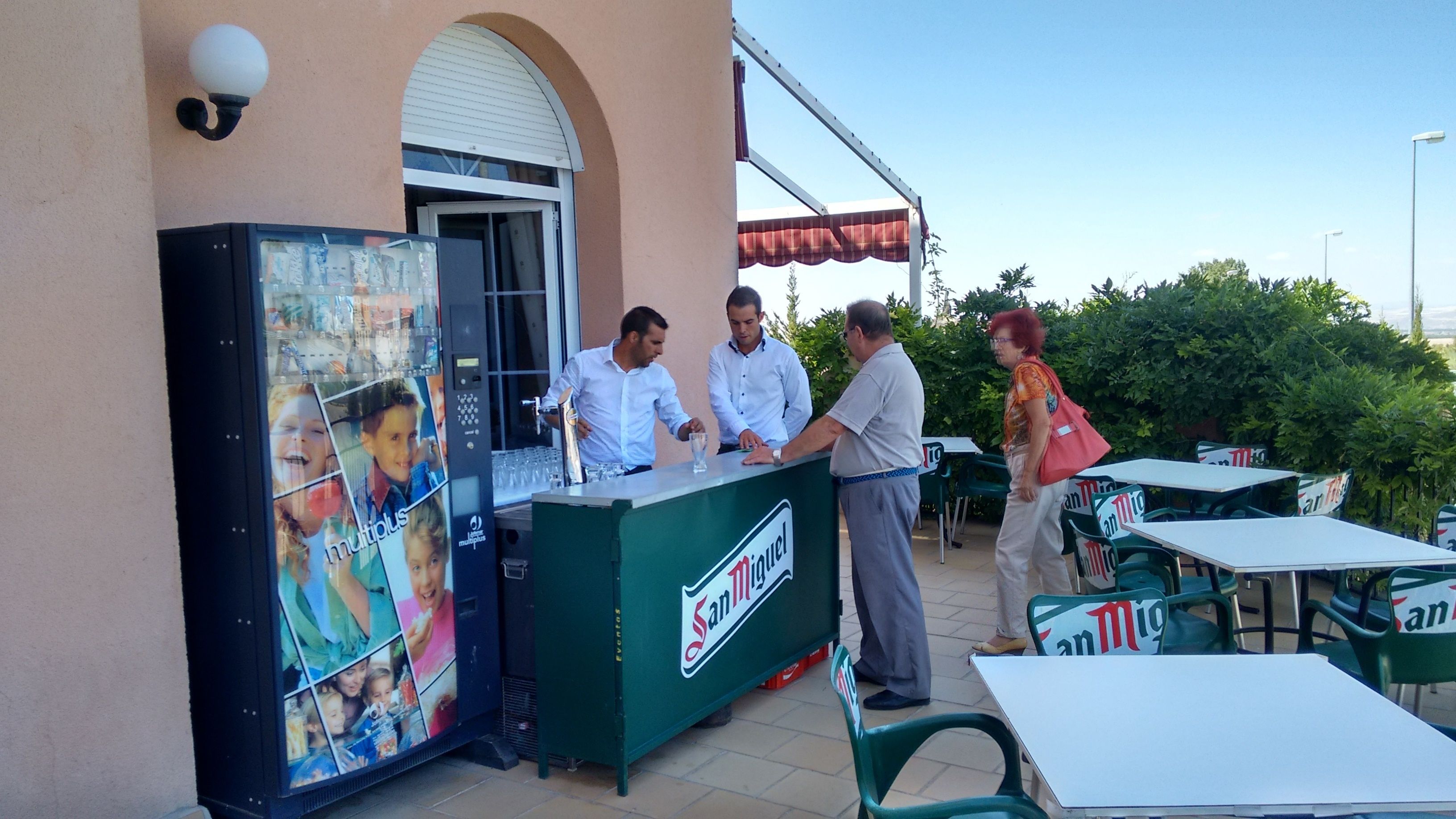 An outdoor bar area with a green counter labeled 'San Miguel', a large advertisement for 'multiplus', and several green tables and chairs.