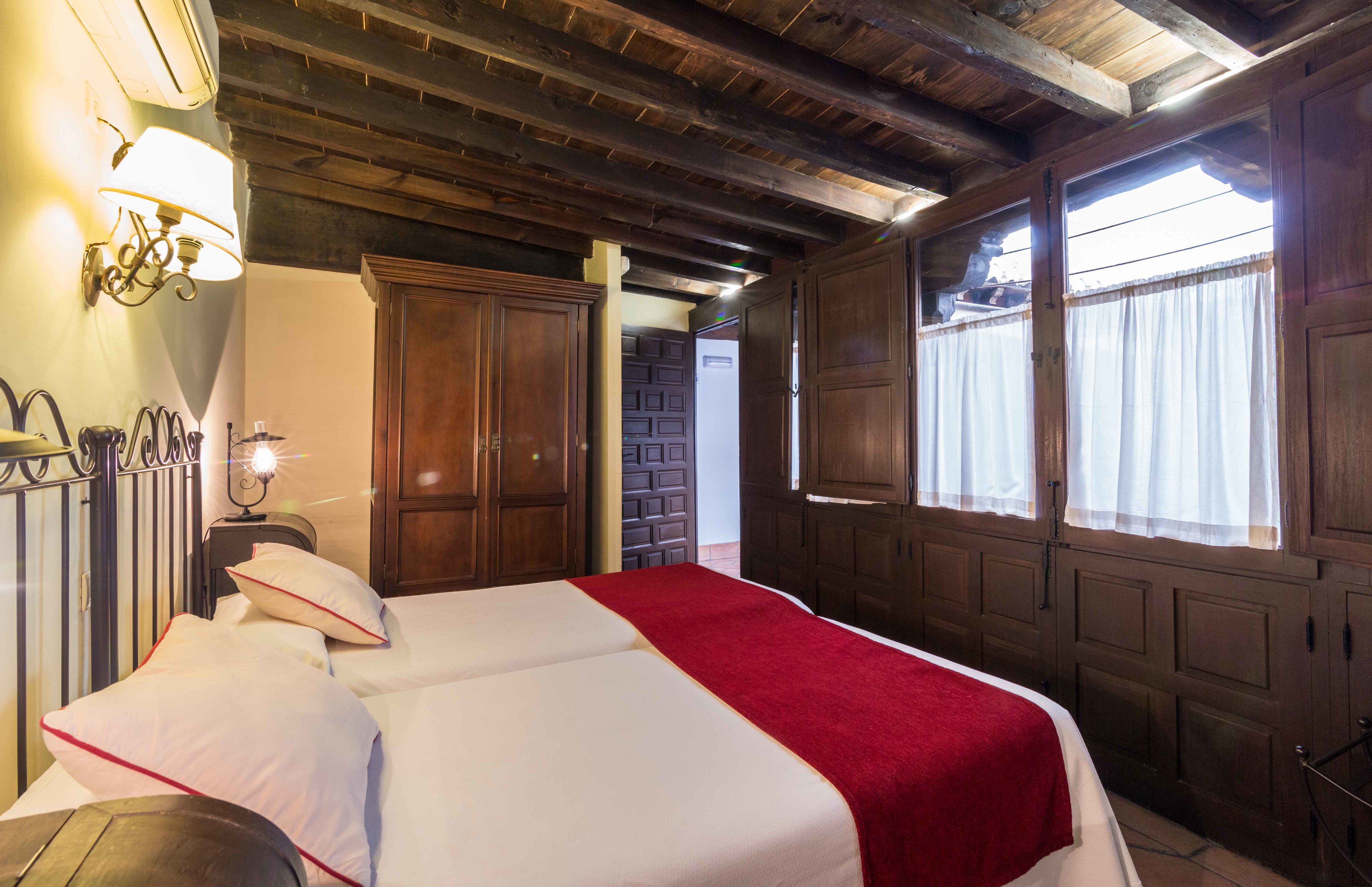 Habitació amb dos llits, bigues de fusta al sostre, una làmpada penjada a la paret i una finestra amb cortines.