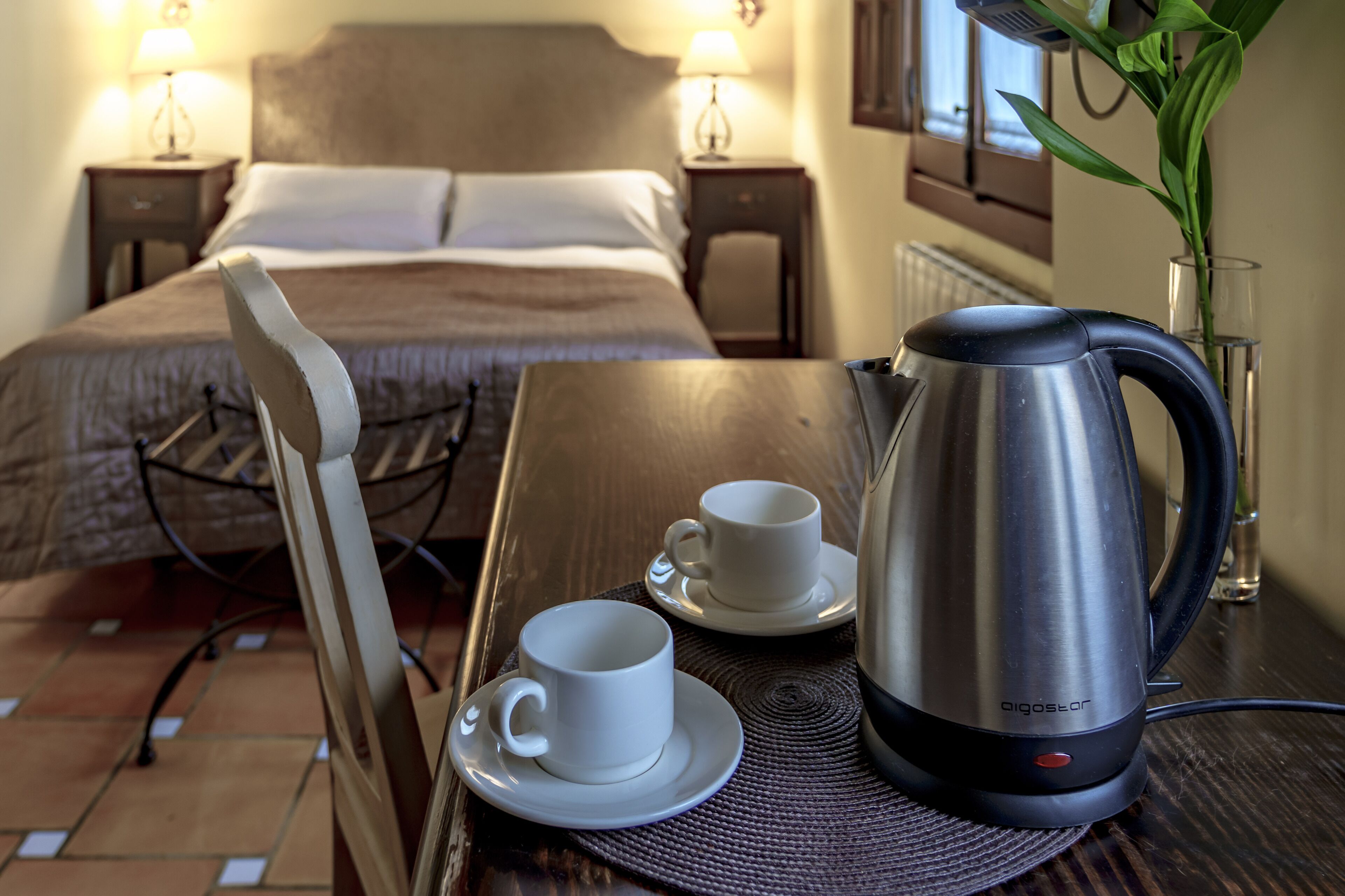A hotel room with a bed, bedside lamps, a table with a kettle, cups, and a chair.