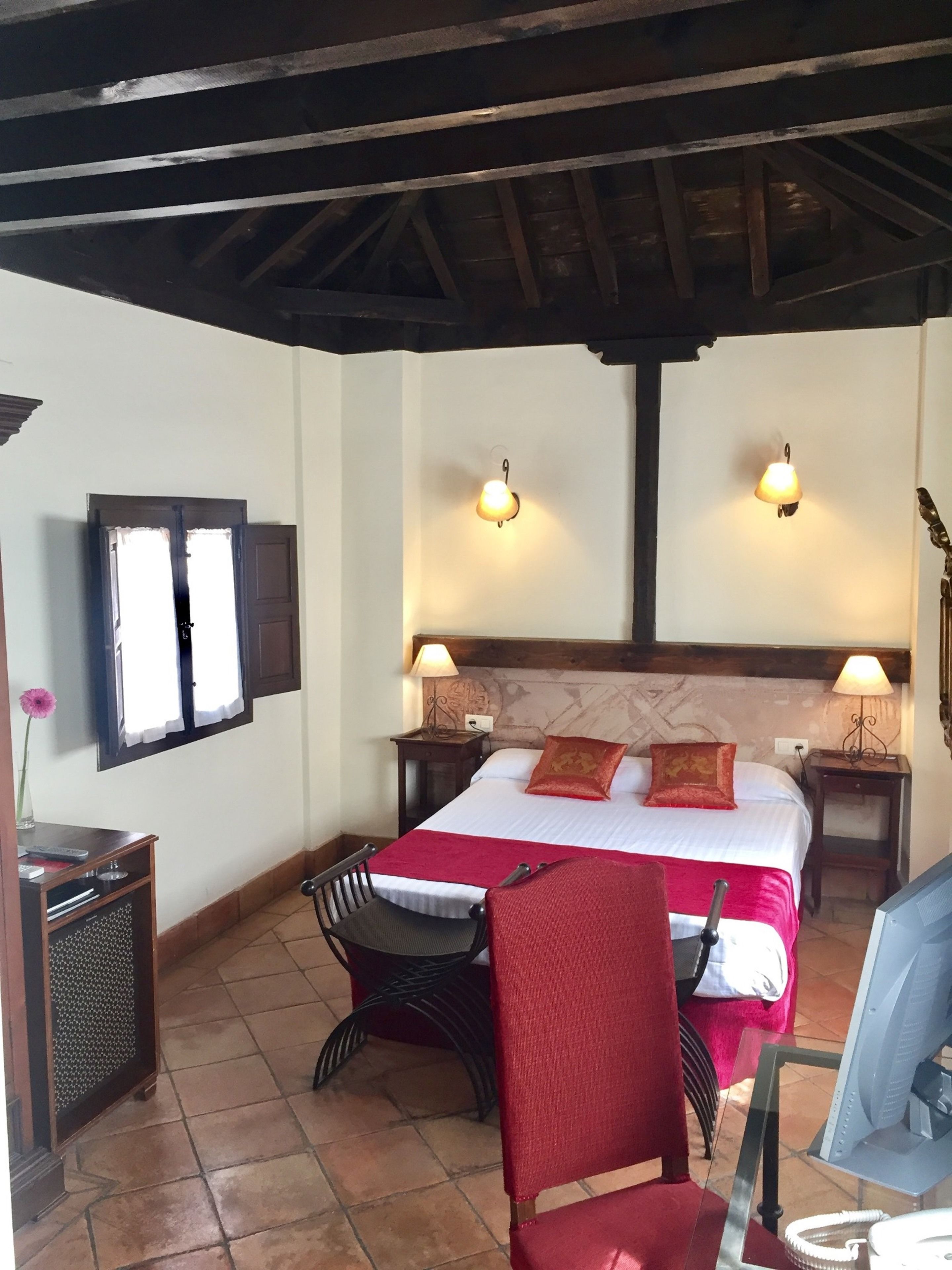 A hotel room with a bed, two bedside lamps, a desk with a chair, a TV, and a small table with a vase.