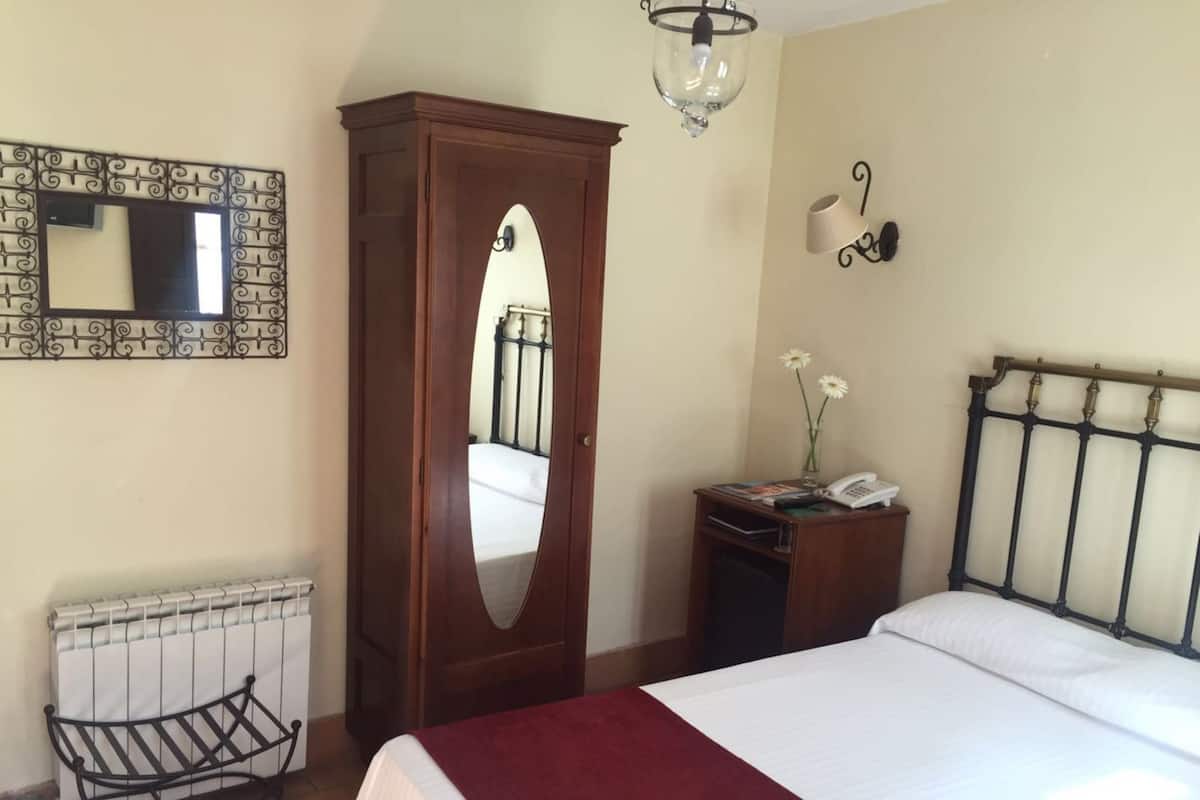 A bedroom with a bed, a wardrobe, a radiator, a mirror, a wall-mounted lamp, and a small table with a phone and flowers.