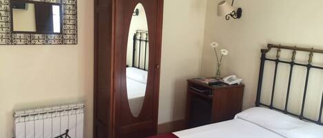 Une chambre à coucher comprenant un lit, une armoire, un radiateur, un miroir, une lampe fixée au mur et une petite table sur laquelle se trouvent un téléphone et des fleurs.