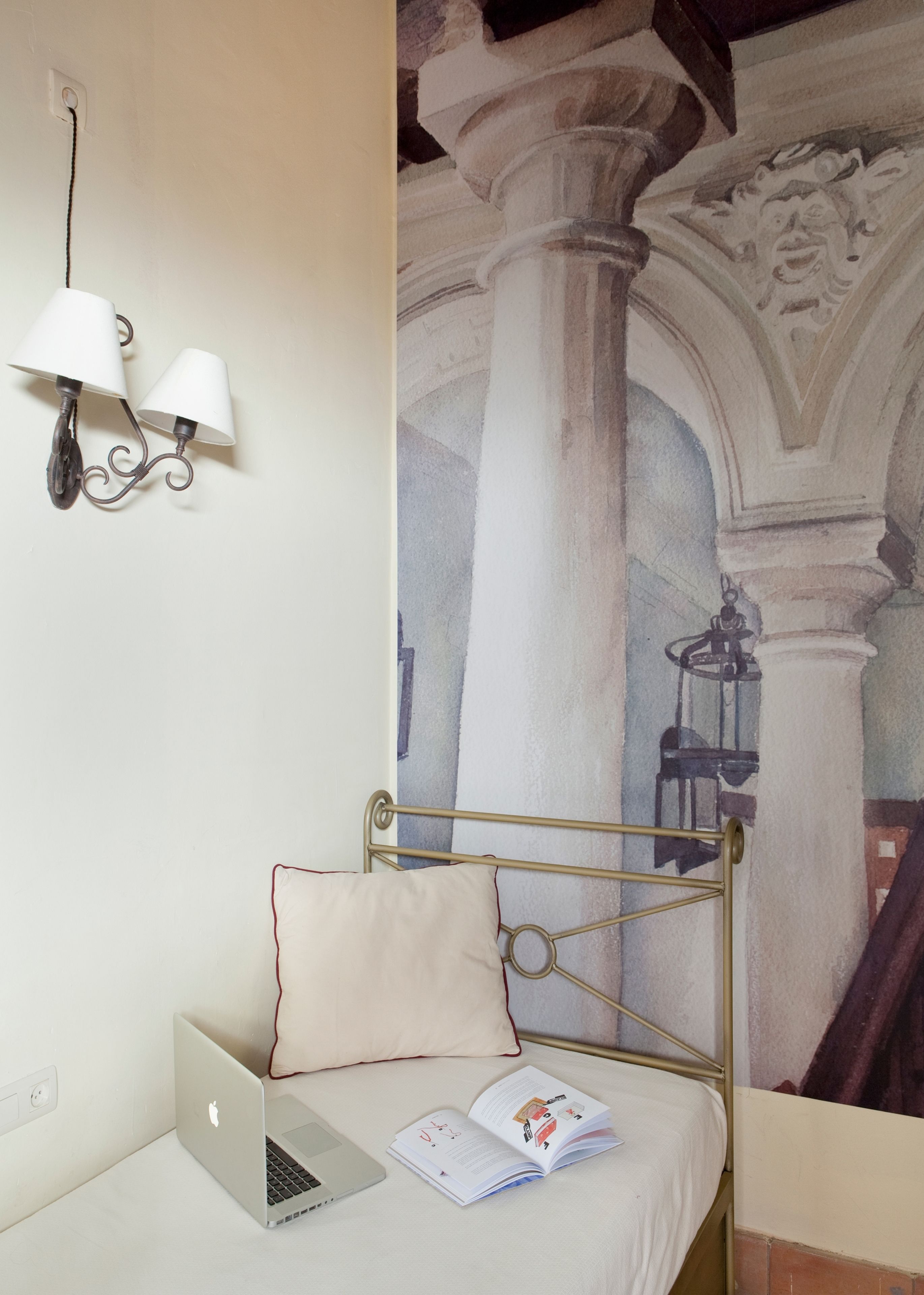 A bed with a laptop and a book, a wall-mounted lamp, and a mural of architectural columns.