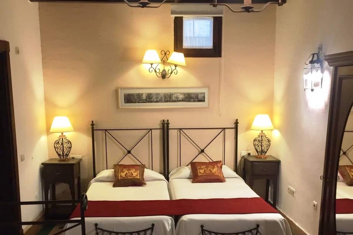 A room with two beds, each with a red bedspread and matching pillows, flanked by nightstands with lamps. There is a framed picture on the wall above the beds, and the room has a tiled floor with a green border.
