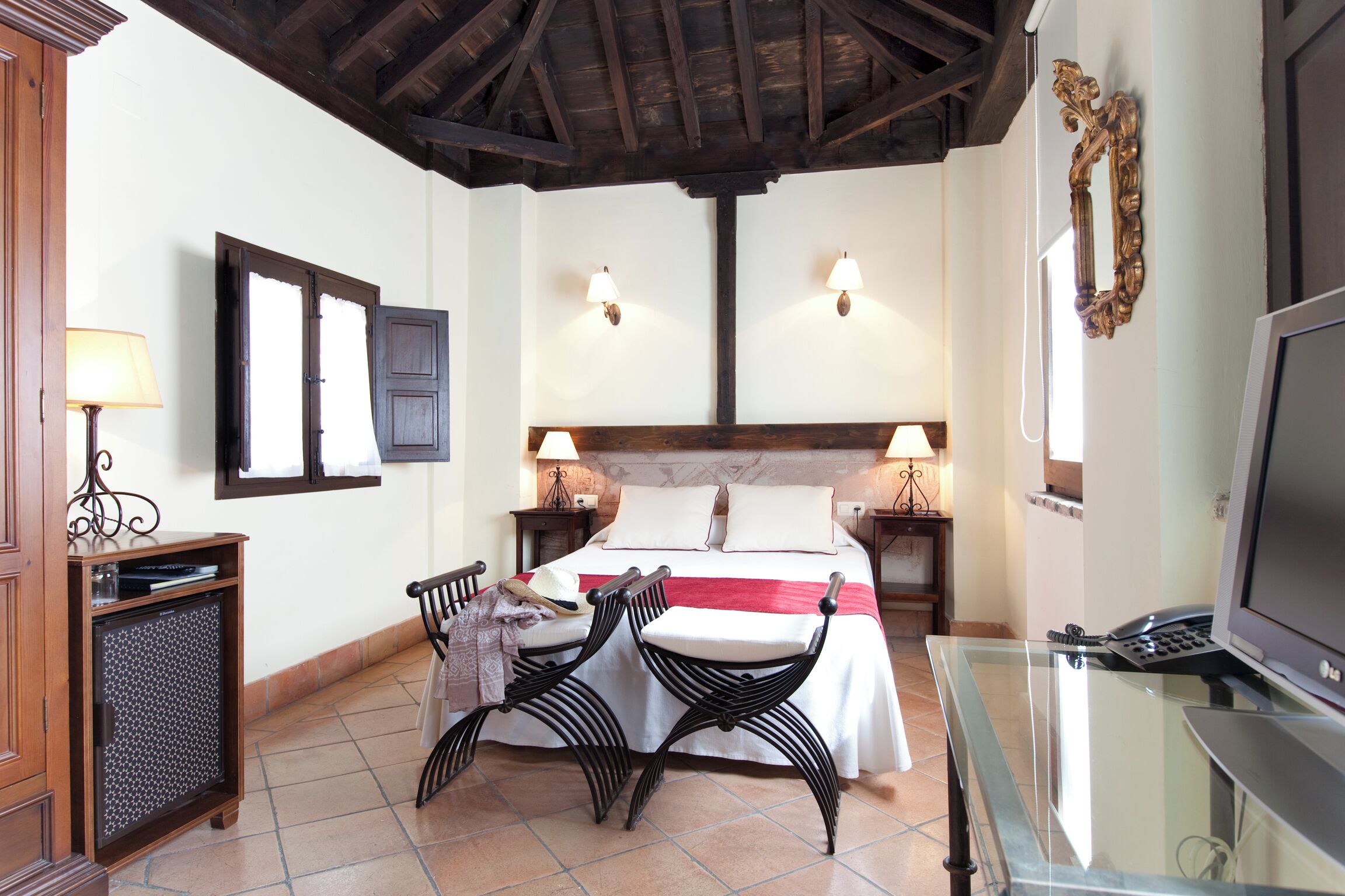 A hotel room with a bed, two bedside tables, a TV, a glass table with chairs, and a wooden cabinet.