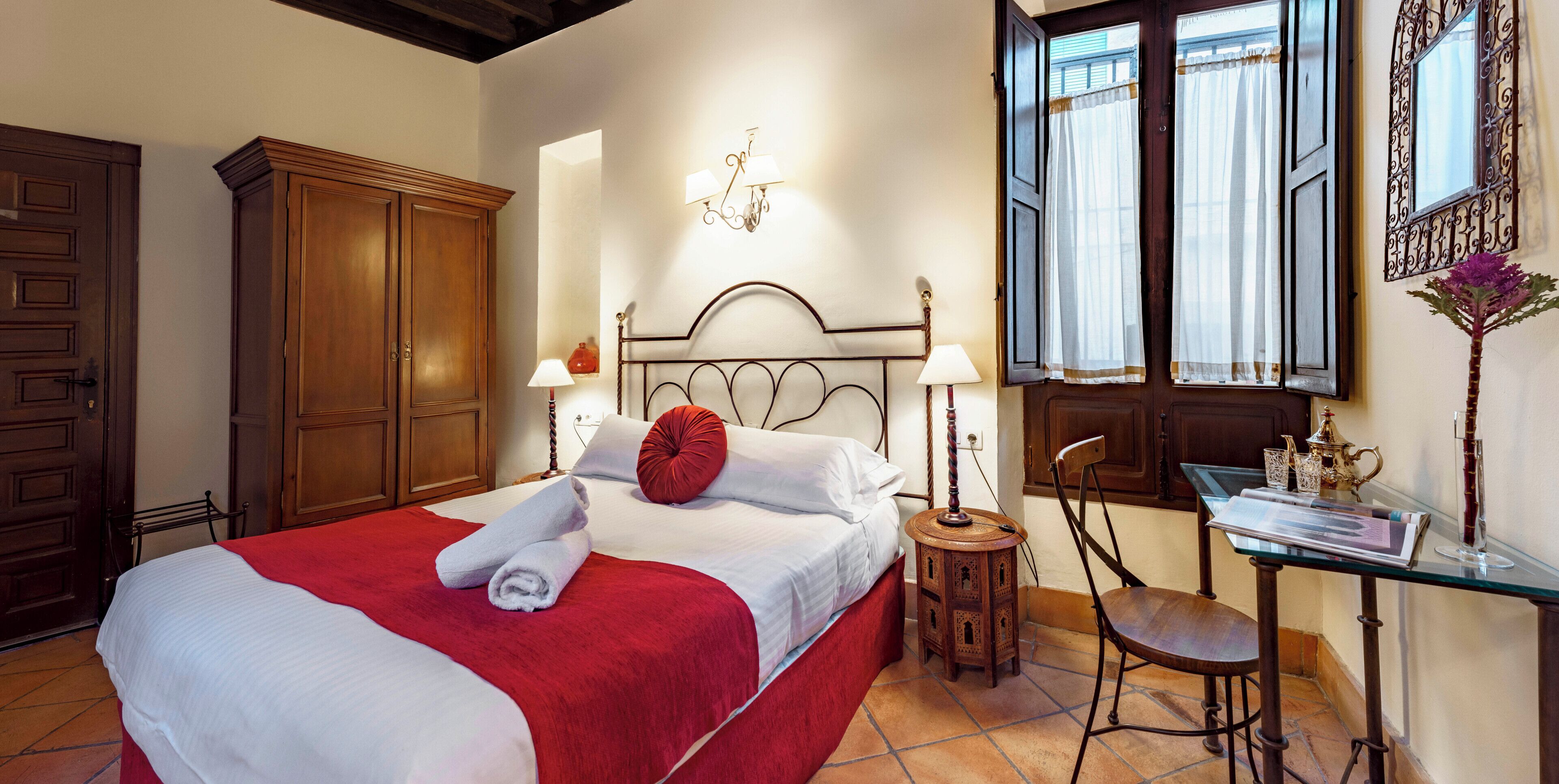A hotel room with a bed, a wooden wardrobe, a desk with a chair, and a window with curtains.