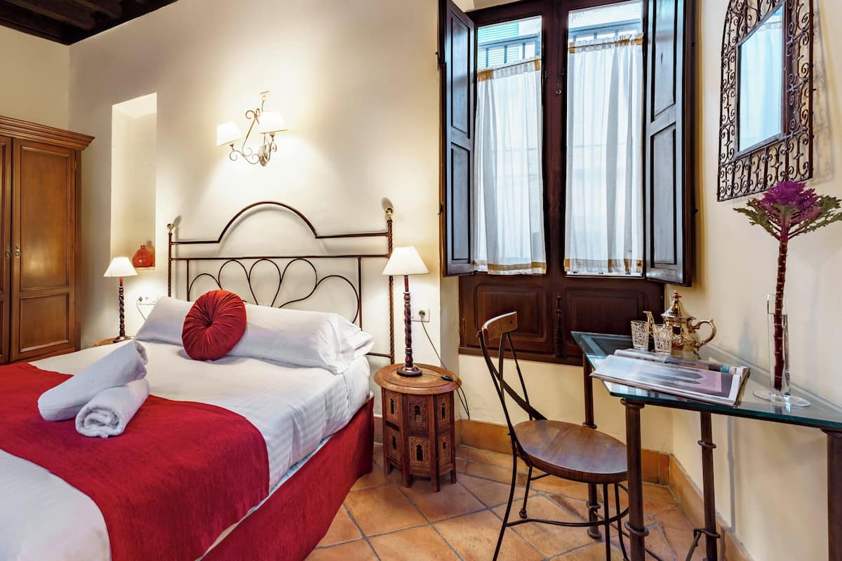 A hotel room with a bed, a wooden wardrobe, a desk with a chair, and a window with curtains.