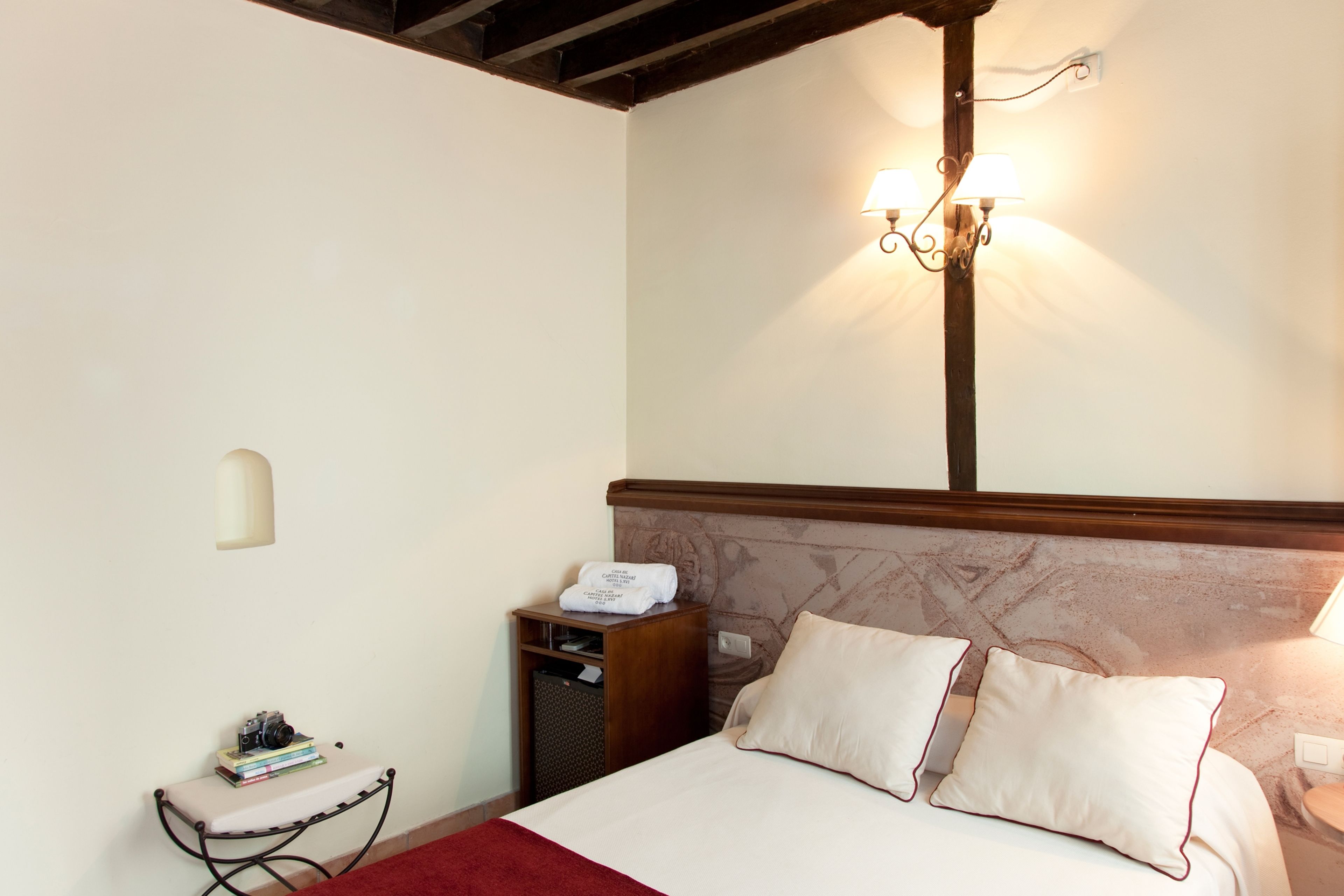 Una habitación de hotel con una cama, mesita de noche, lámpara y una pequeña mesa cubierta con un paño rojo.