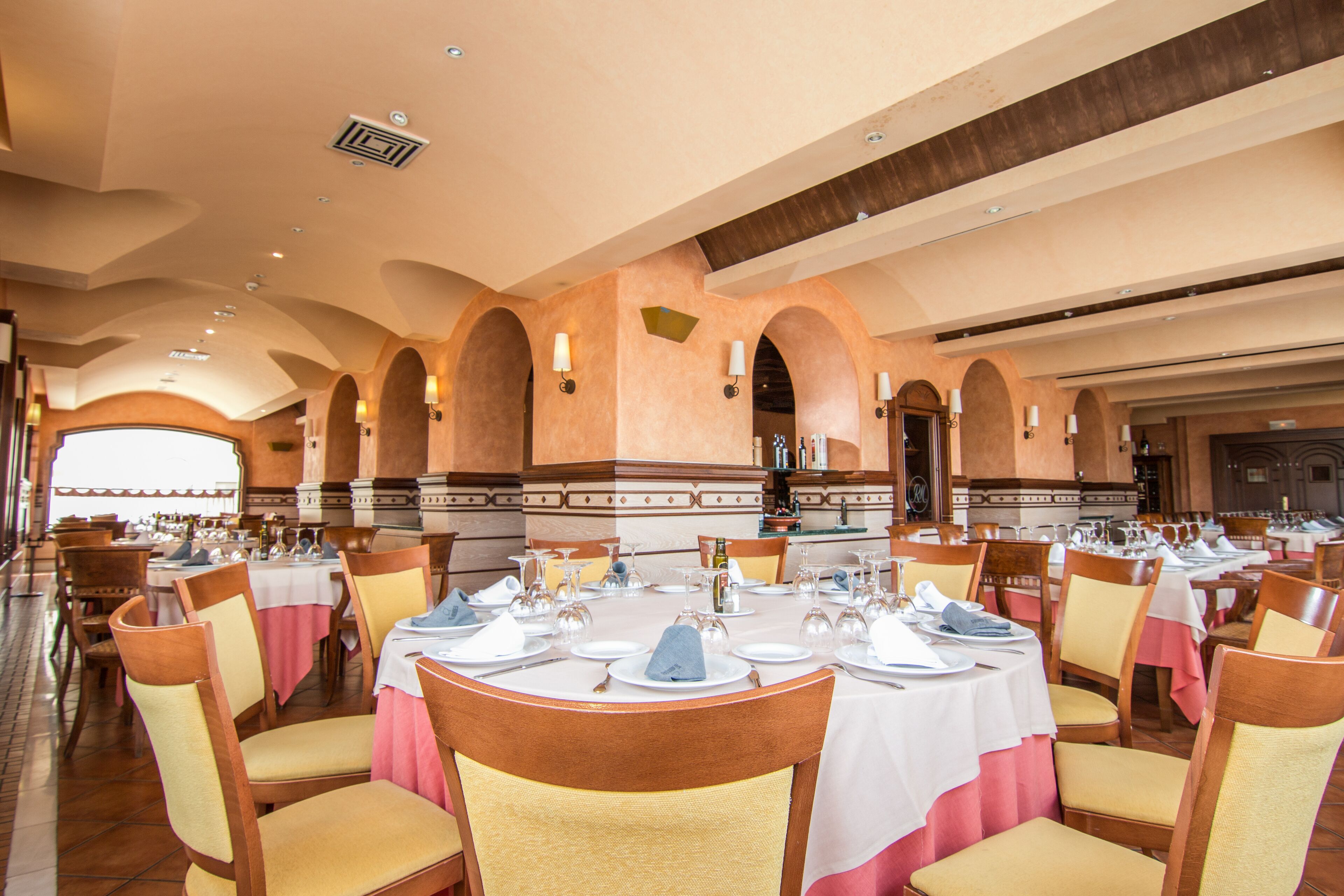 A large dining hall with multiple round tables, each set with white tablecloths, chairs, and dinnerware.