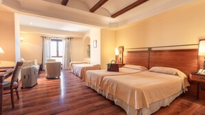 Habitación de hotel con tres camas, piso de madera, un escritorio con silla, una maleta y una lámpara.