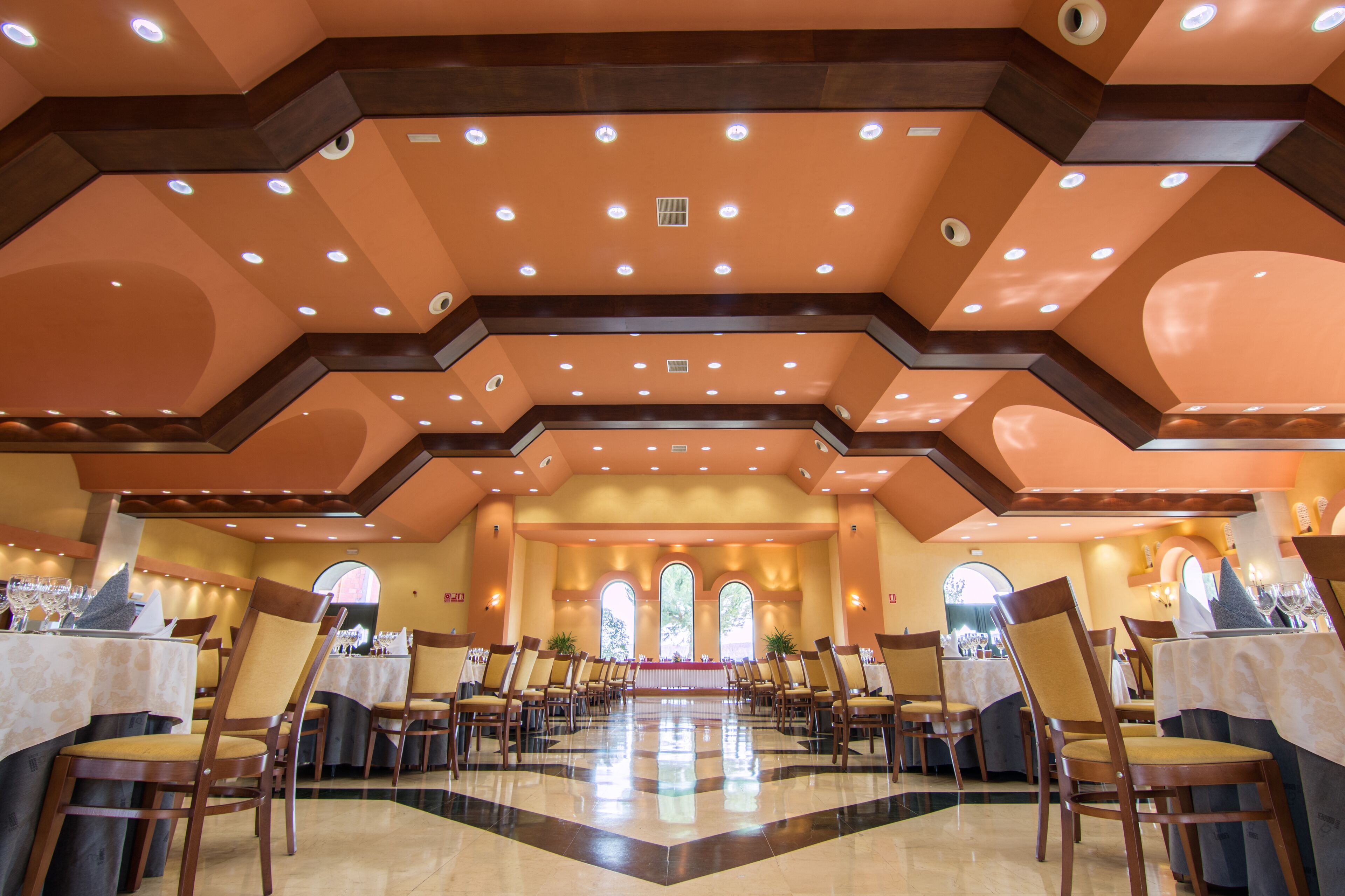 A spacious dining hall with high ceilings, multiple chandeliers, and a central aisle leading to a large window.