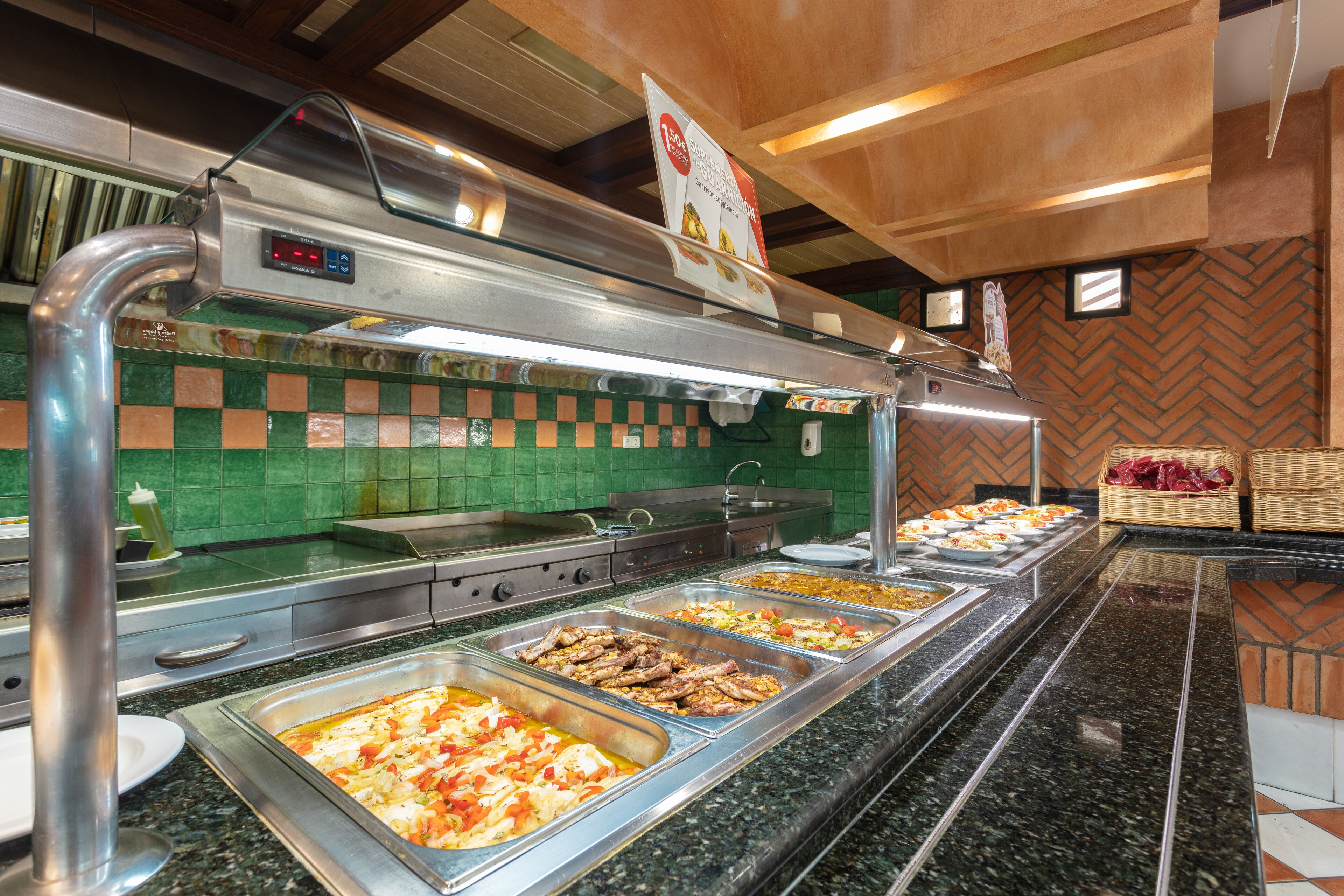 Un bufé con diversas opciones de comida, un lavamanos de acero inoxidable y un salpicadero de azulejos verdes.