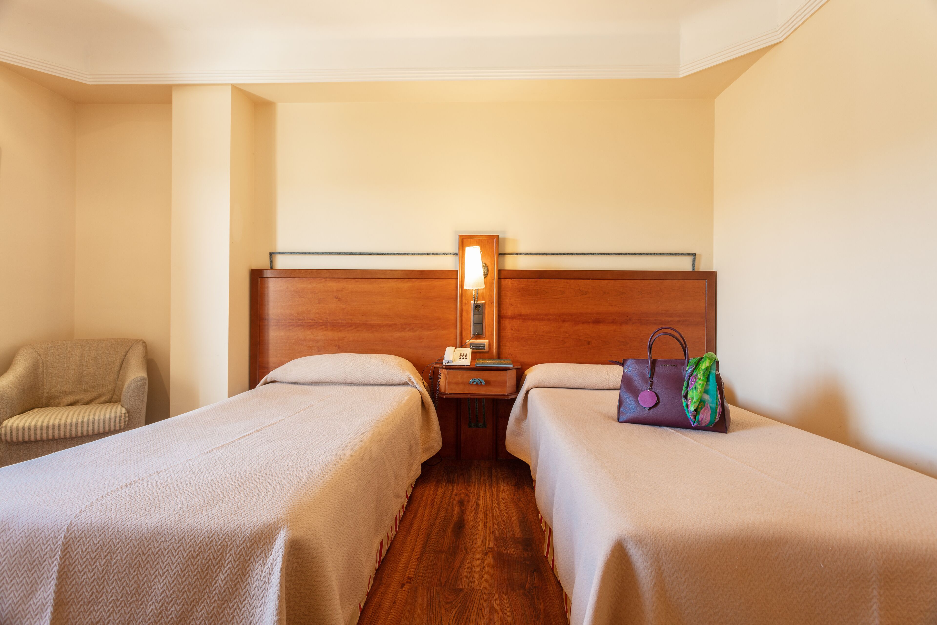 A hotel room with two single beds, a wooden headboard, a nightstand with a lamp, a chair, and a handbag on the bed.