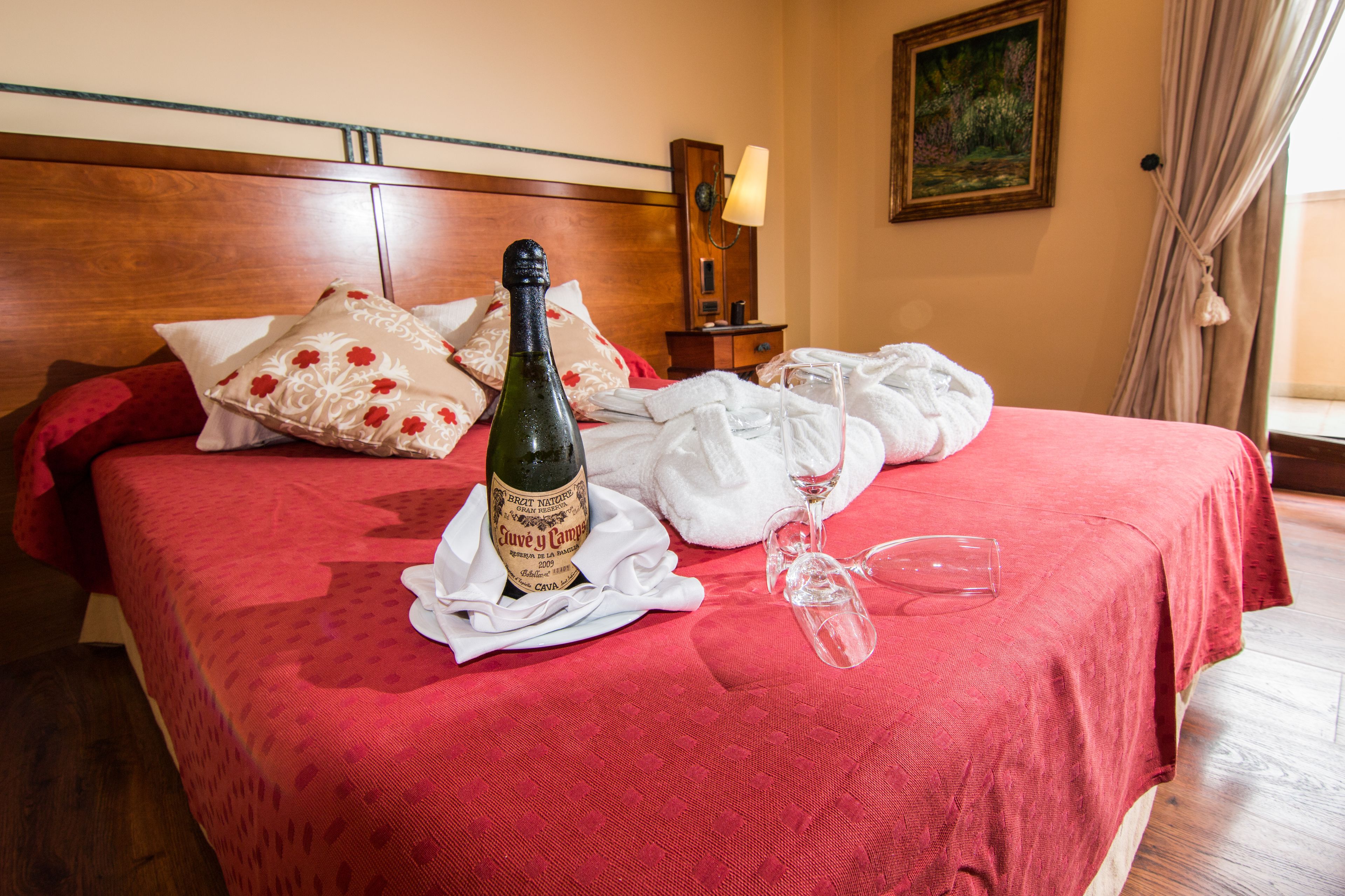 Una cama bien tendida con un edredón rojo, una botella de vino y una copa de vino encima.