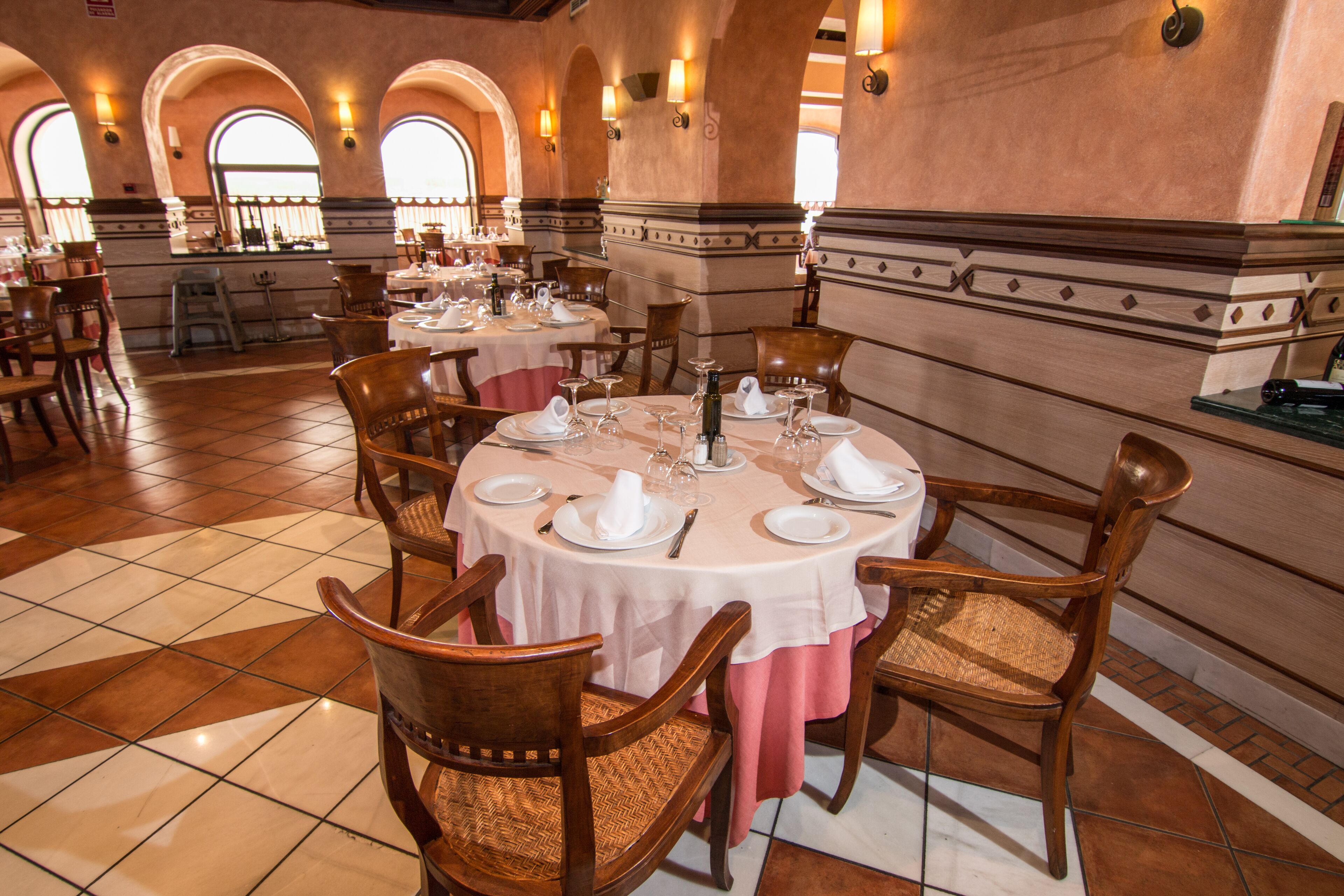 Comedor con mesas redondas dispuestas para varios comensales, con sillas de madera y manteles blancos.