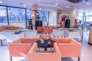 Un área de descanso moderna con asientos naranja y gris, una mesa central con bebidas y ventanas amplias que ofrecen vistas al exterior.