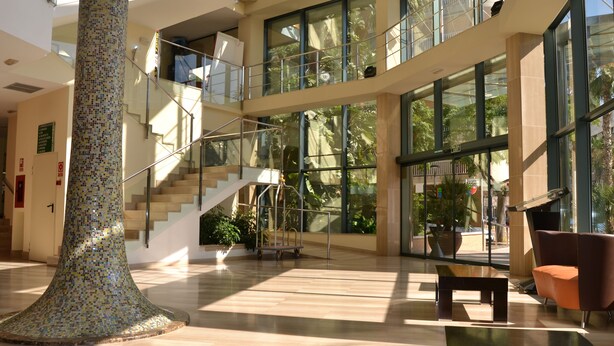 A spacious lobby with a large mosaic column, a staircase, and a seating area with a modern chair. - Hotel California Palace (Salou)