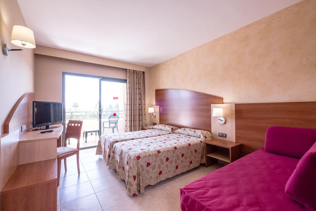 A hotel room with two beds, a desk with a TV, a chair, a balcony with a view, and a red sofa.