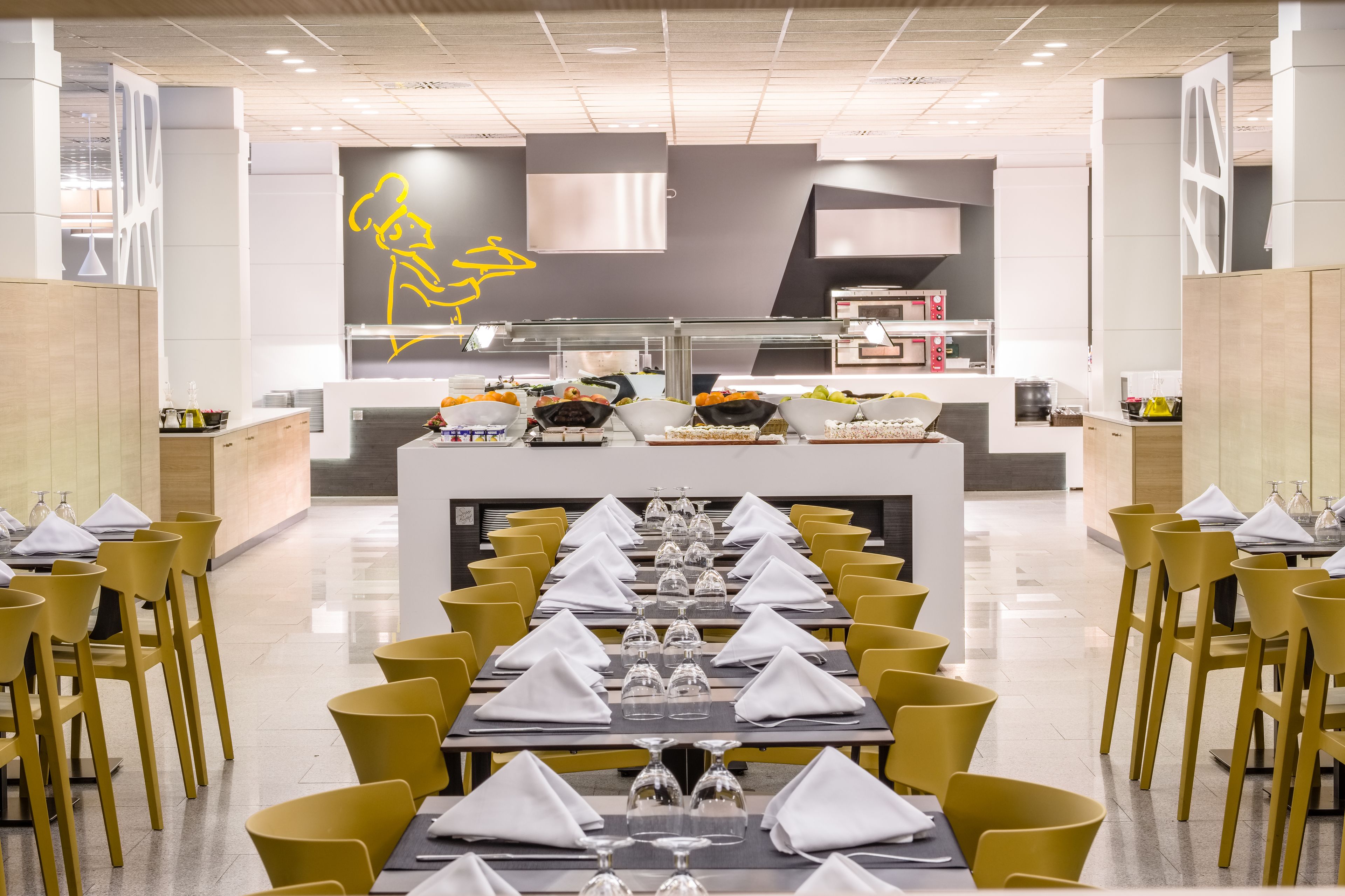 Een modern restaurant met gele stoelen en tafels, een centrale toonbank met etalages en een minimalistische inrichting.