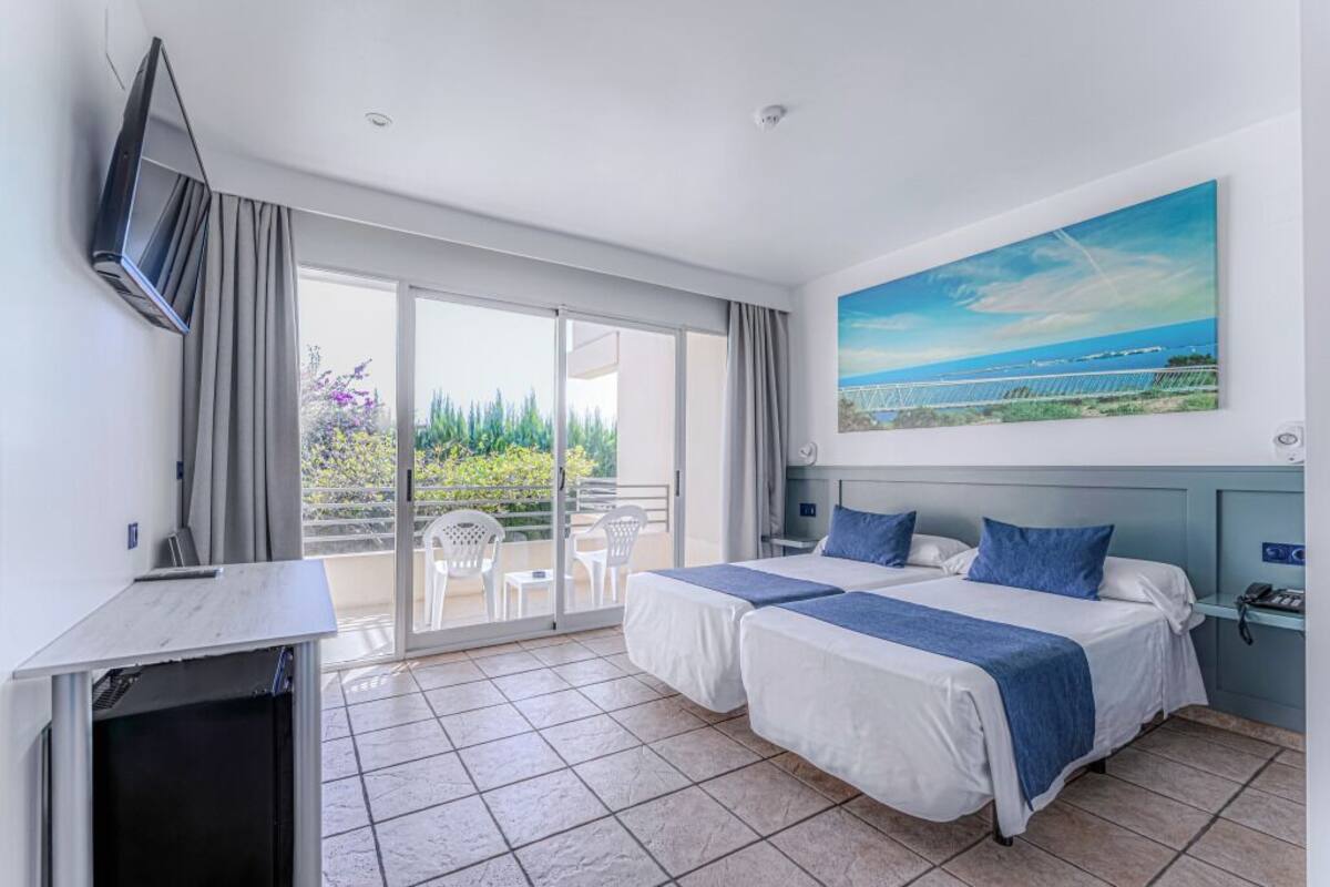A hotel room with two beds, a desk, a TV, and a balcony with a view of the sea.