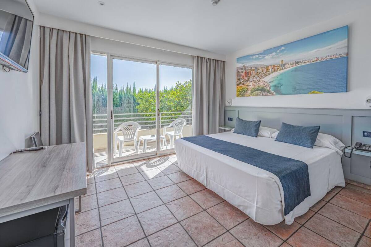 A hotel room with a large bed, a desk, a TV, and a balcony with a cityscape view.