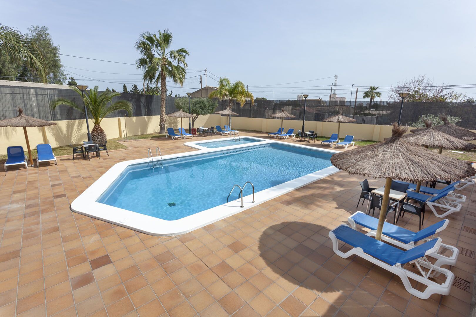 Una piscina rectangular con borde blanco, rodeada de sillas reclinables azules y sombrillas de paja.