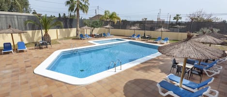 A rectangular swimming pool with a white edge, surrounded by blue lounge chairs and thatched umbrellas.