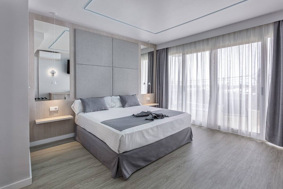 A modern bedroom with a large bed, a bedside table, a mirror, and a window with curtains.