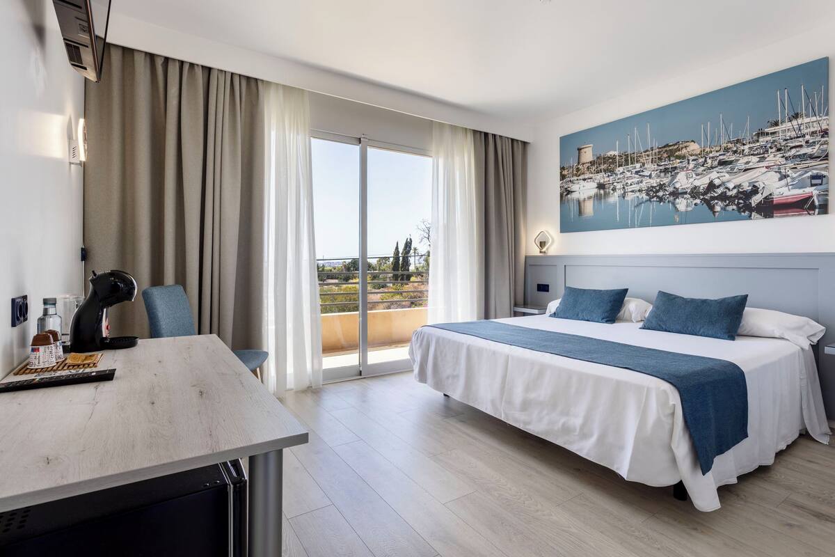 A hotel room with a bed, a desk, a chair, a large window with curtains, and a painting of a marina.