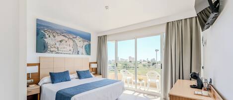 A hotel room with a large bed, a desk, a TV mounted on the wall, and a balcony with a view of the city.