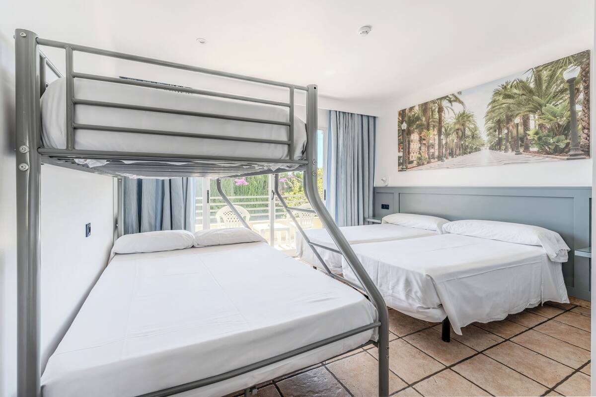 A dormitory room with bunk beds, a painting of a tropical scene, and a view of greenery outside.