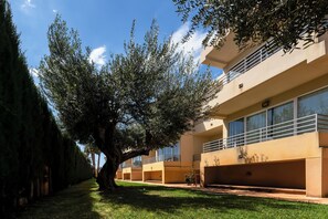 Il s’agit d’un bâtiment à l’architecture moderne, caractérisé par un toit plat et de grandes fenêtres. Un arbre se trouve au premier plan, offrant de l’ombre, et un ciel bleu clair se déploie au-dessus.