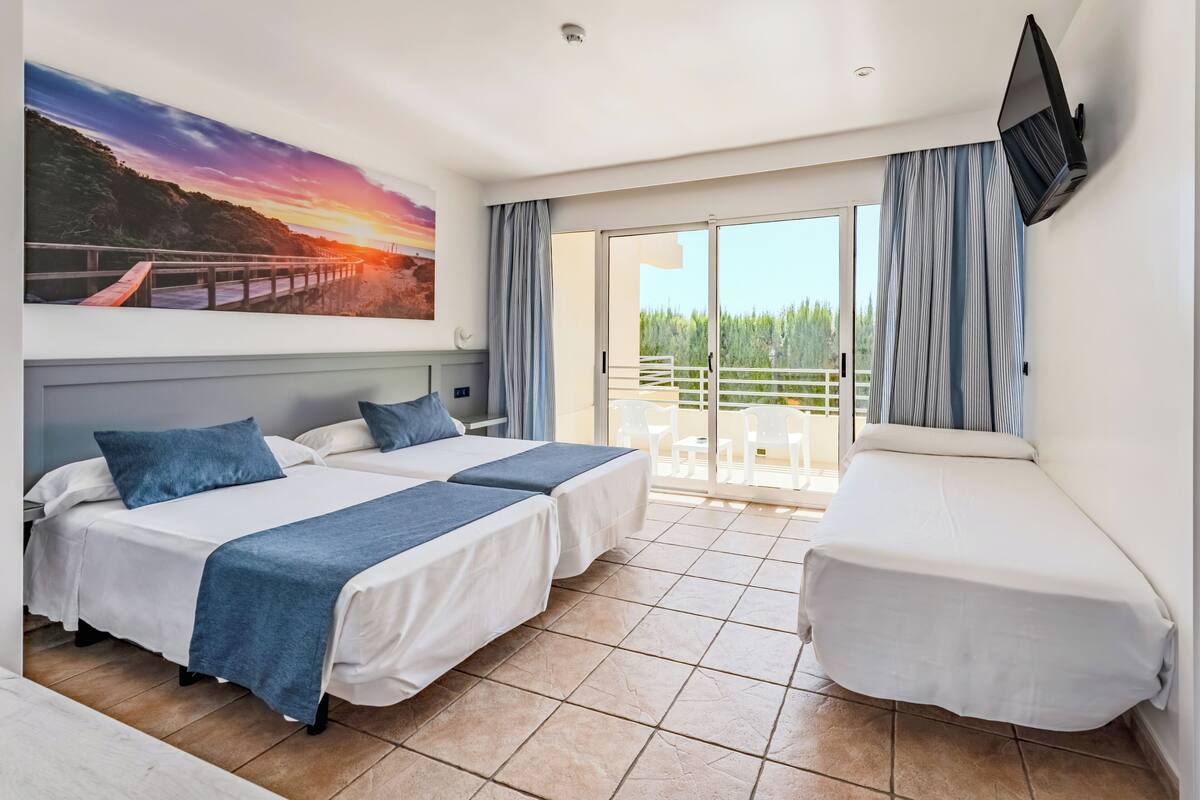 A hotel room with two beds, a TV, a balcony with a view of the forest, and a sunset painting on the wall.